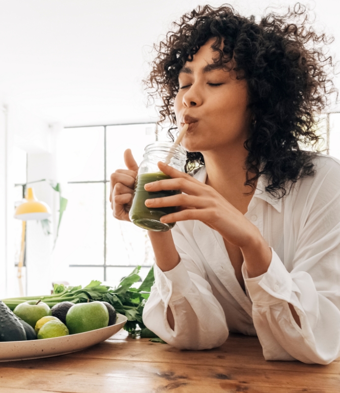 Frau mit dunklen Locken, die grünen Smoothie trinkt, symbolisiert die richtige Ernährung bei Akne-Behandlung