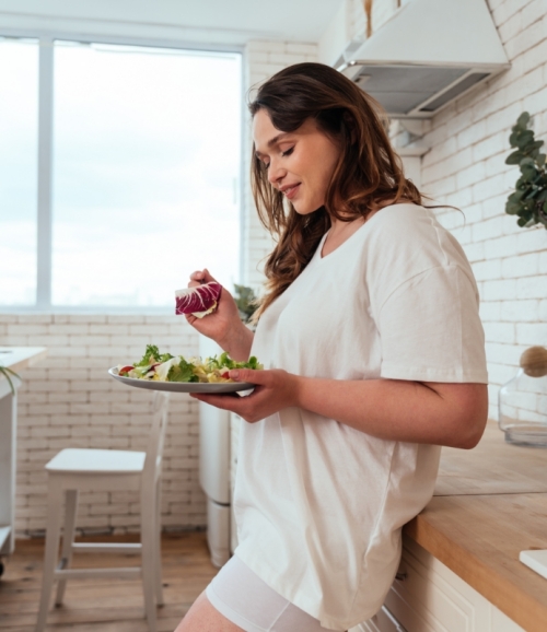 Frau, die an Küchentresen lehnt und Salat isst, steht für die Vorbereitung auf Liposuktion