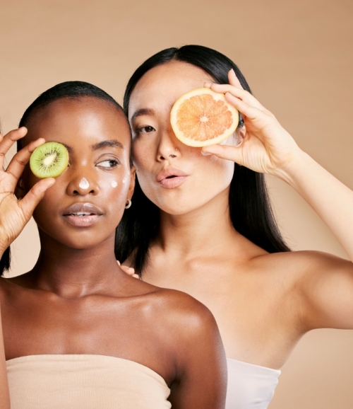 Zwei junge Frauen mit Kiwi- bzw. Grapefruitscheibe vor Auge symbolisieren Ernährungsberatung als Teil der Rosacea-Behandlung in Elbklinik